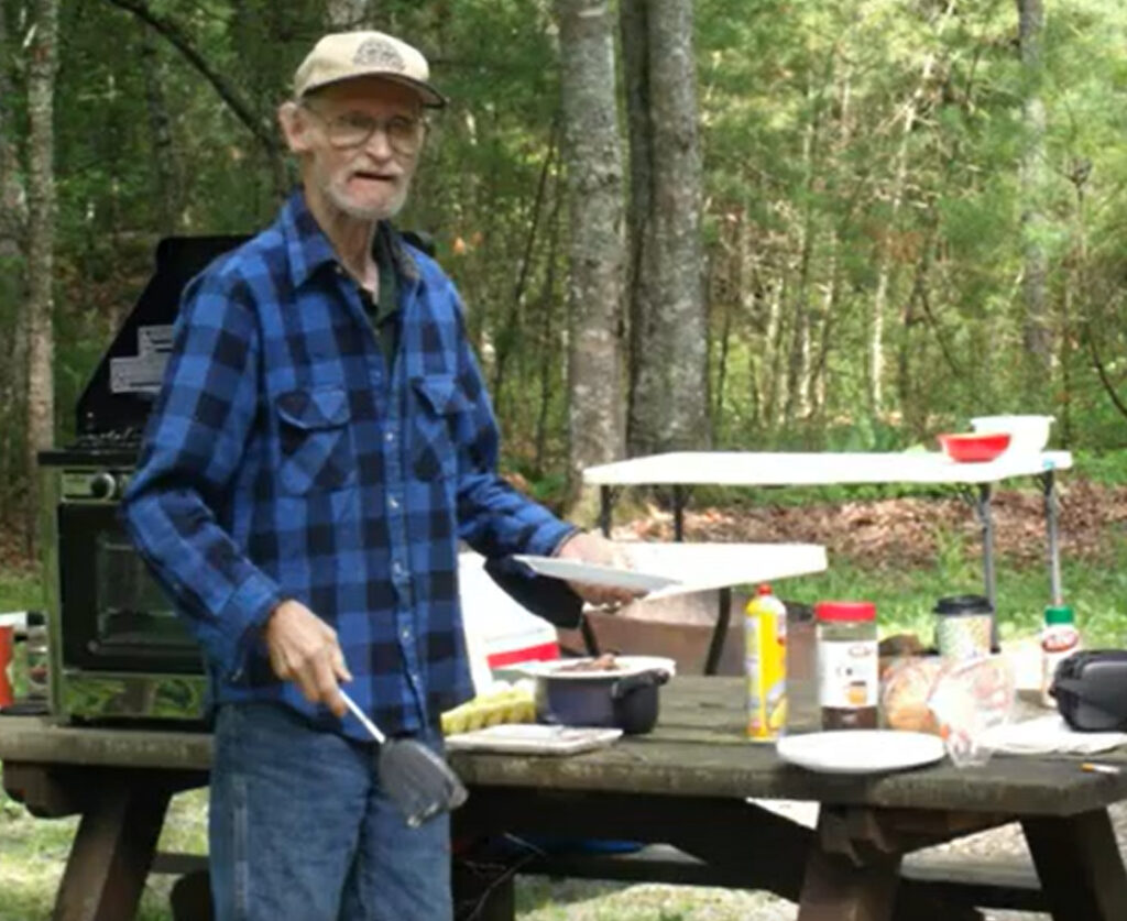Elderly man in flannel shirt cooking outdoors at a picnic table in the woods